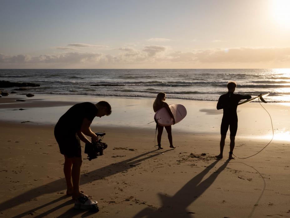 Camera operator at the beach with two surfers, sunset